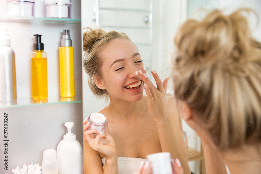 Young beautiful woman applying moisturising cream on skin in bathroom. Standing in towel with little jar of moisturizer, looking in the mirror, laughing and having fun. Morning skincare routine. Young beautiful woman applying moisturising cream on skin in bathroom. Standing in towel with little jar of moisturizer, looking in the mirror, laughing and having fun. Morning skincare routine.