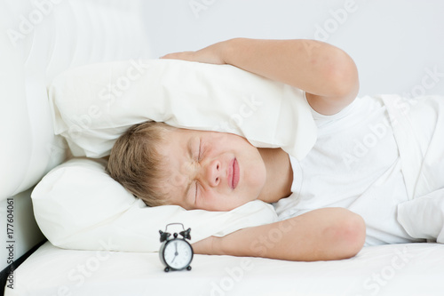 the boy clamps his ears with a pillow, not wanting to hear alarm clock