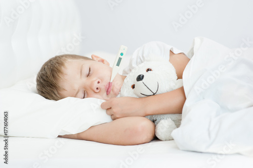 Sick boy with thermometer in mouth lying in bed and hugging a toy bear