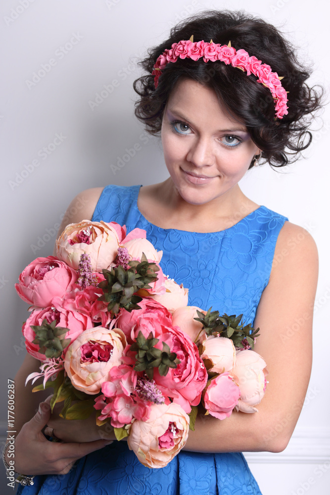 Smiling young brunette woman with clean skin, make-up and and flower wreath in her hair on grey background with bouquet of flowers in hands, looks slyly Smiling young brunette woman with clean skin, make-up and and flower wreath in her hair on grey background with bouquet of flowers in hands, looks slyly