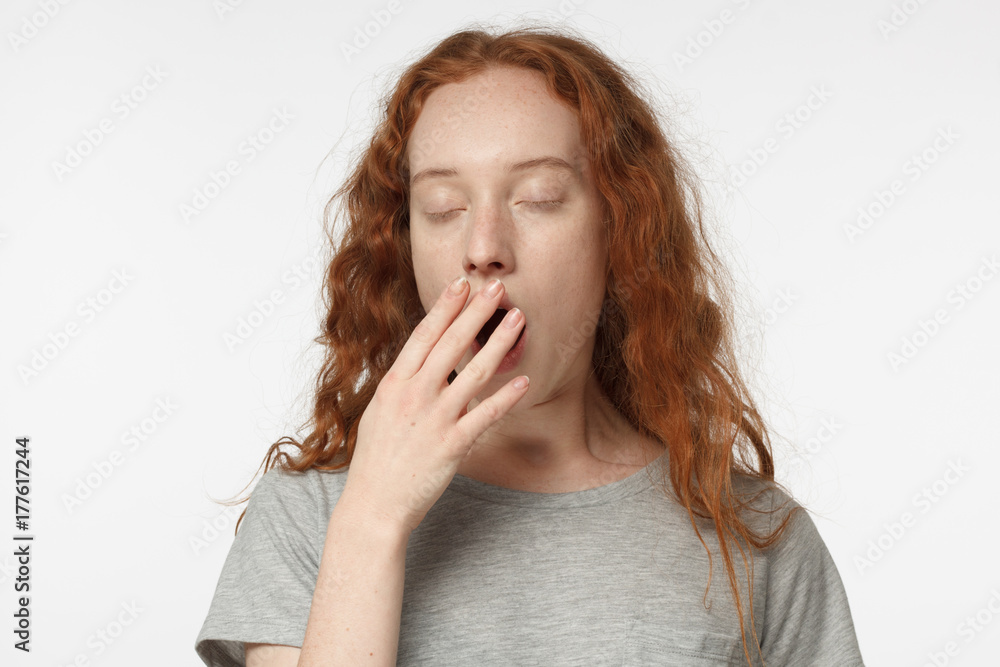 Sleepy redhead girl yawning, waking up, closing her mouth with hand, isolated on gray background Sleepy redhead girl yawning, waking up, closing her mouth with hand, isolated on gray background