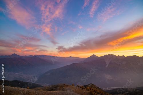 The Italian French Alps at sunset. Colorful sky over the majestic mountain peaks, dry barren terrain and green valleys. Sunburst and backlight expansive view from above.