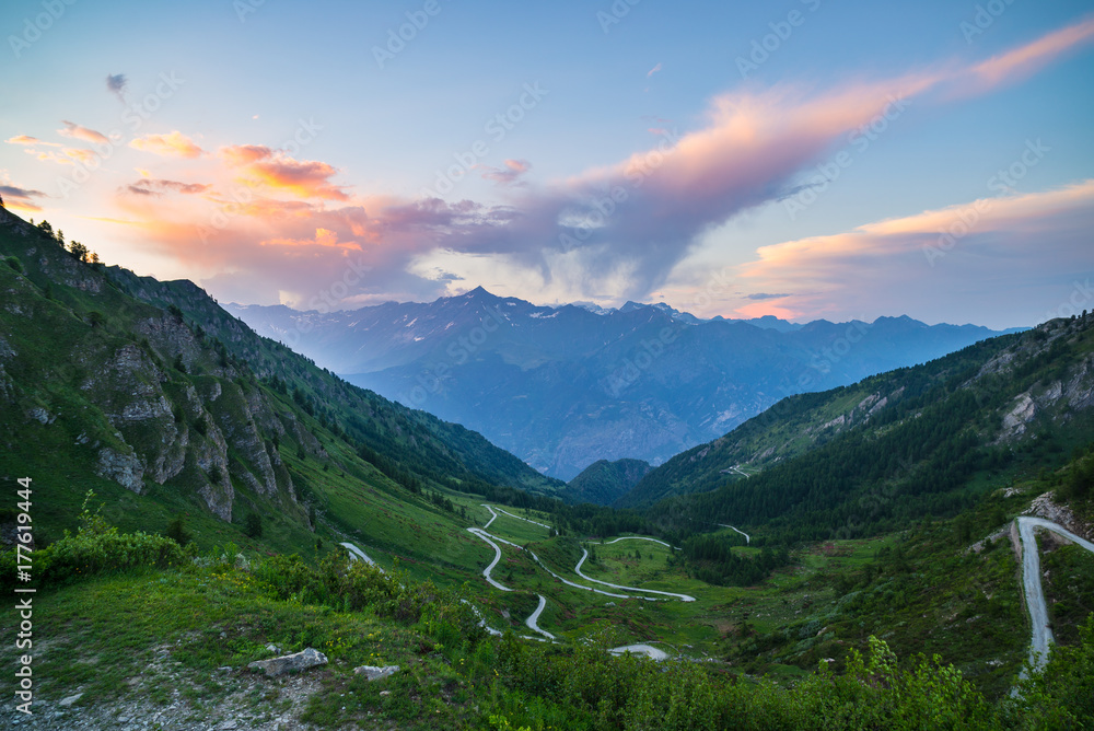 Mountain road leading to high mountain pass on the Italian Alps.. Expasive view at sunset, colorful dramatic sky, adventure road trip in summer. Mountain road leading to high mountain pass on the Italian Alps.. Expasive view at sunset, colorful dramatic sky, adventure road trip in summer.