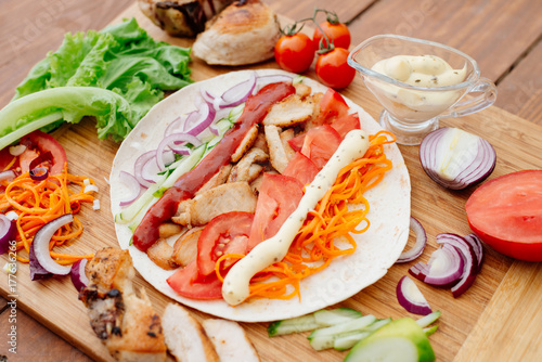 The process of preparing chiken shawarma. Ingredients sliced lie on the kitchen board. Fast food. Eastern food. Dishes of oriental cuisine lying on pita bread and decorated with greens and vegetables.
