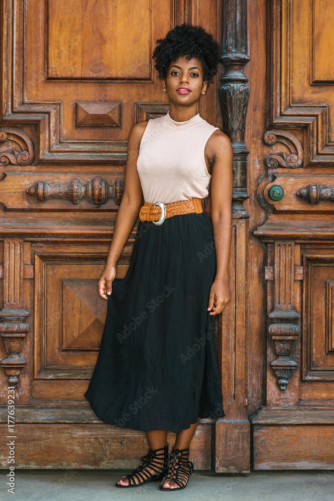 African American Businesswoman working in New York. Young black female  teacher with afro hairstyle wearing sleeveless light color top, black skirt,  strappy sandals, standing by vintage office door.. Photos | Adobe Stock