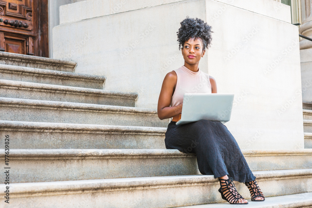 Way to Success. Young African American woman with afro hairstyle wearing sleeveless top, black skirt, sandals, sitting on stairs of vintage office building in New York, working on laptop computer..