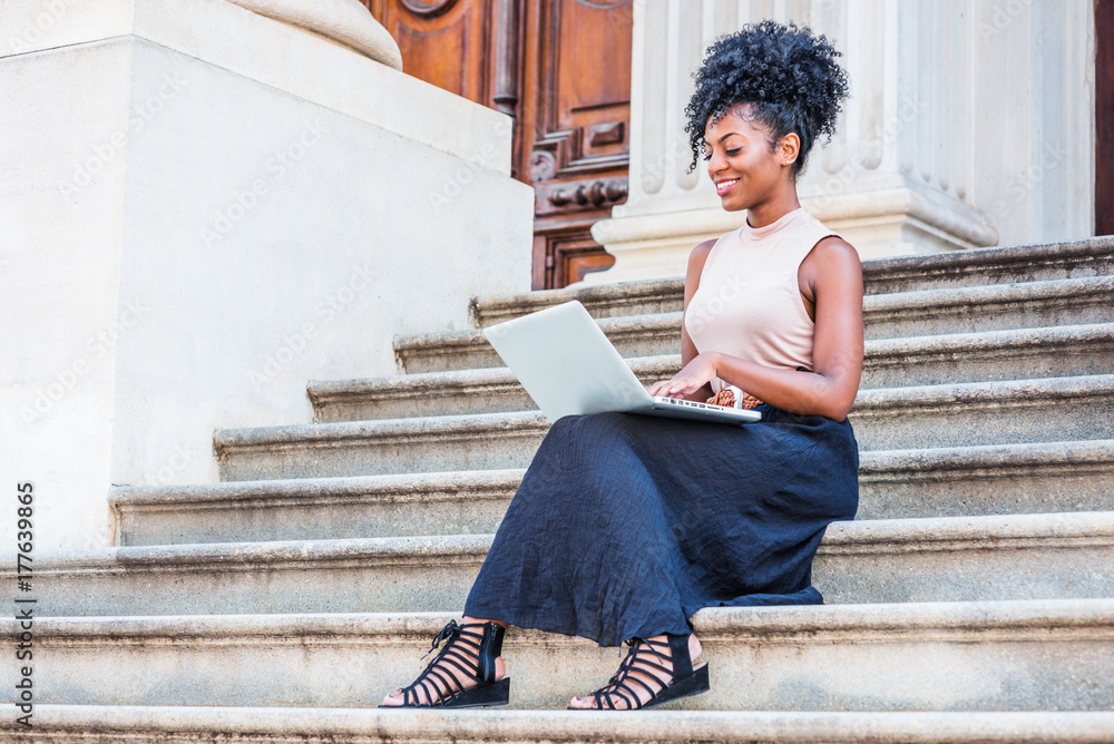 Way to Success. Young African American woman with afro hairstyle wearing sleeveless top, black skirt, sandals, sitting on stairs of vintage office building in New York, working on laptop computer..