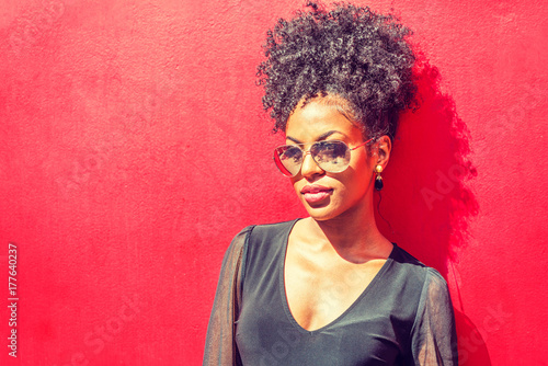 Portrait of Young African American Woman in New York. Young black woman with afro hairstyle wearing long sleeve mesh sheer shirt, sunglasses, standing against red background under sun..