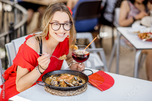 Young woman in red dress eating sea Paella, traditional Valencian rice dish, sitting outdoors at the restaurant in Valencia, Spain