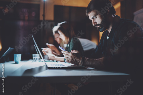 Bearded young coworker working at night office.Man using contemporary laptop and modern smartphone.Horizontal.Blurred background. Flares and reflections effect. photo