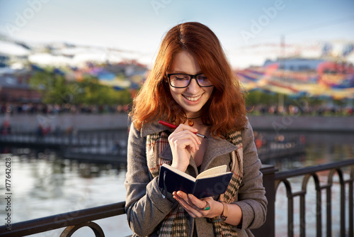 Smiling cheerful girl makes note in notebook, planning how to organize holiday for weekend. young attractive red-haired girl with freckles makes notes in a notebook on background of sunny park. photo