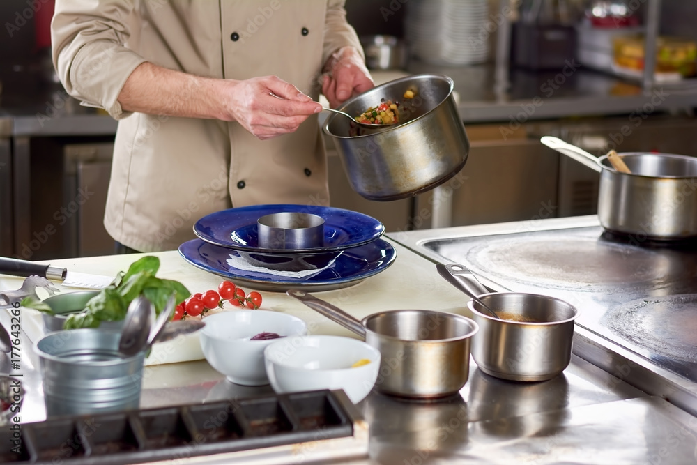 Chef put out with spoon stewing vegetables. Cropped image of male chef going putting garnishing into stainless bowl. Food preparation at professional kitchen. Chef put out with spoon stewing vegetables. Cropped image of male chef going putting garnishing into stainless bowl. Food preparation at professional kitchen.