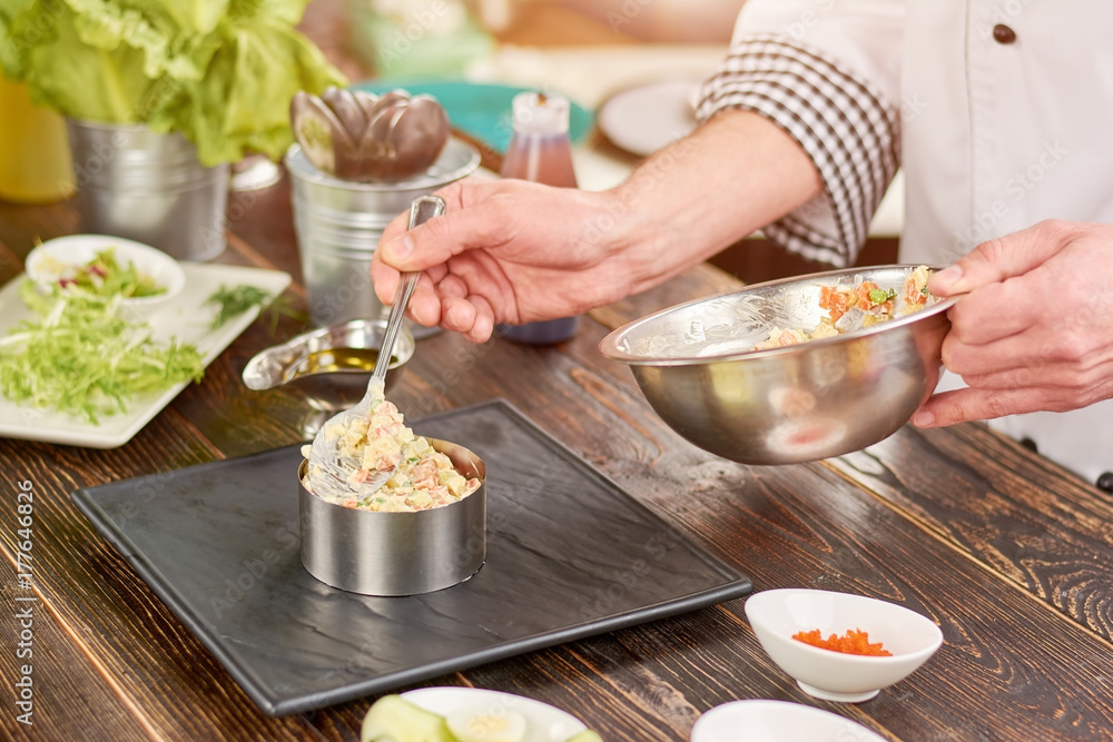 Chef making an appetizing salad. Chef hand putting salad with spoon in metal ring. Cook preparing olivier salad at professional kitchen. Chef making an appetizing salad. Chef hand putting salad with spoon in metal ring. Cook preparing olivier salad at professional kitchen.