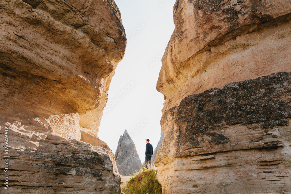The man in dark clothes and baseball cap stands between beautiful rocks and admires the landscape in Cappadocia in Turkey