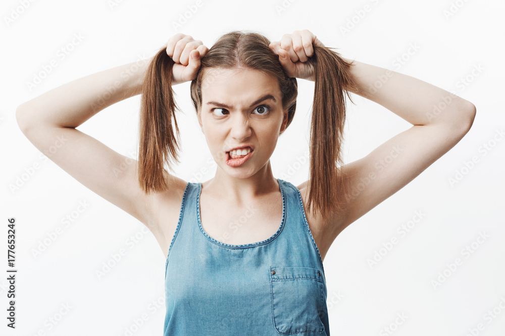 Close up of funny beautiful european female student with brown hair in blue jeans shirt holding hair with both hands, mowing eyes, showing teeth, looking in camera with awkward expression, having fun. Close up of funny beautiful european female student with brown hair in blue jeans shirt holding hair with both hands, mowing eyes, showing teeth, looking in camera with awkward expression, having fun.