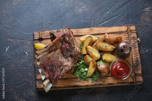 Wooden serving tray with roasted lamb meat on bone and potato wedges, high angle view on a dark brown stone surface