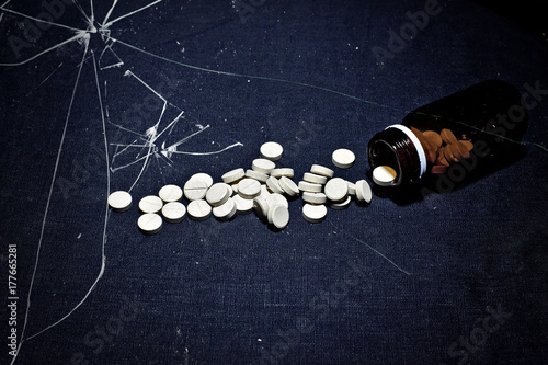 White pills on a table cloth spilled from an opaque glass bottle, seen under a broken glass.
