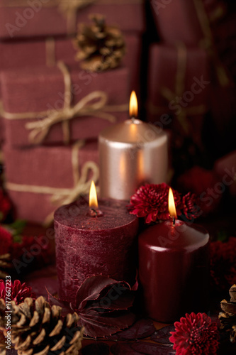 Three candles of crimson and pink color on the background of gift boxes with cones  leaves and daisies.