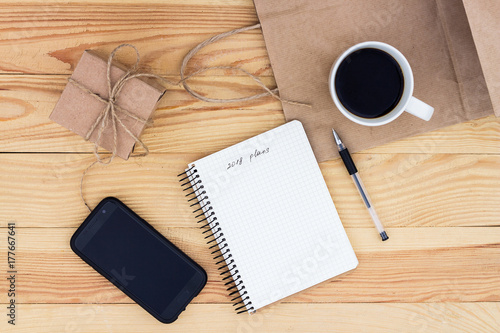 A notebook with an inscription 2018 plans  laying on wooden table. It is surrounded by   cup of coffee   present  black pen  phone  and a paper bag.