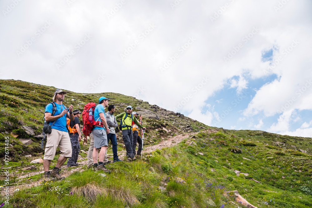 A group of hikers with backpacks and tracking sticks rest and stands in the mountains listening to their guide