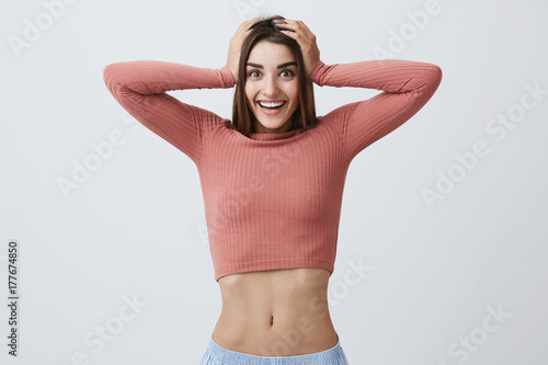 Close up portrait of beautiful joyful young caucaian girl with dark long hair in pink stylish top and blue jeans smiling with teeth, holding head with hands, looking in camera with happy and excited