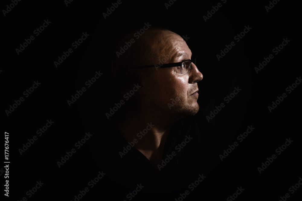 Side profile of a white middle aged man with spectacles or glass in shadow against a black background looking up towards the light Side profile of a white middle aged man with spectacles or glass in shadow against a black background looking up towards the light