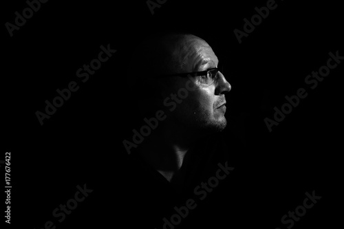 Side profile of a white middle aged man with spectacles or glass in shadow against a black background looking up towards the light