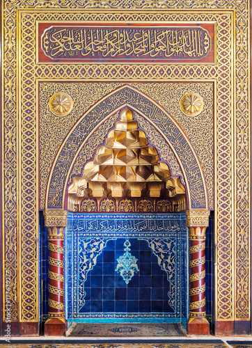 Golden ornate arched mihrab (niche) with floral pattern, blue Turkish ceramic tiles and arabic calligraphy at the public mosque of The Manial Palace of Prince Mohammed Ali Tewfik, Cairo, Egypt photo