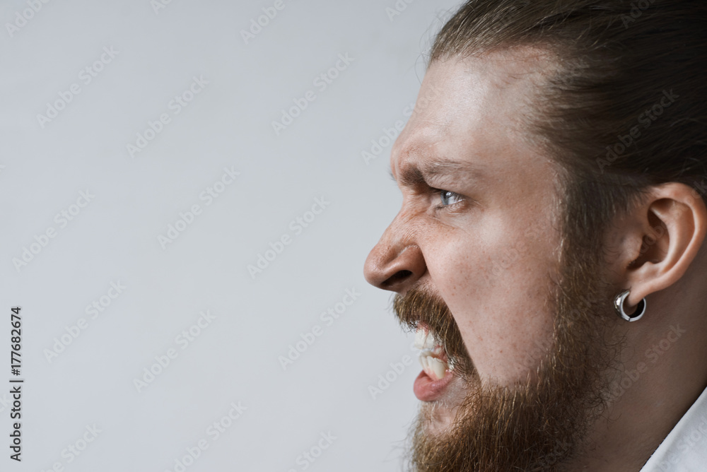 Obraz premium Close up profile of aggressive mad young man with earring and bushy beard clenching teeth in frenzy or rage, can't control his feelings, freaking out because of stress, failure or problems at work