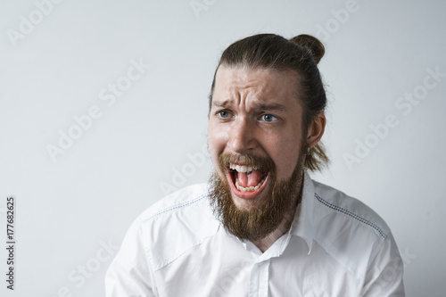 Studio shot of outraged furious young man with thick beard and hair knot screaming with anger and rage, frowning and opening mouth widely while having argument, dispute or fight, loosing his temper © Anatoliy Karlyuk