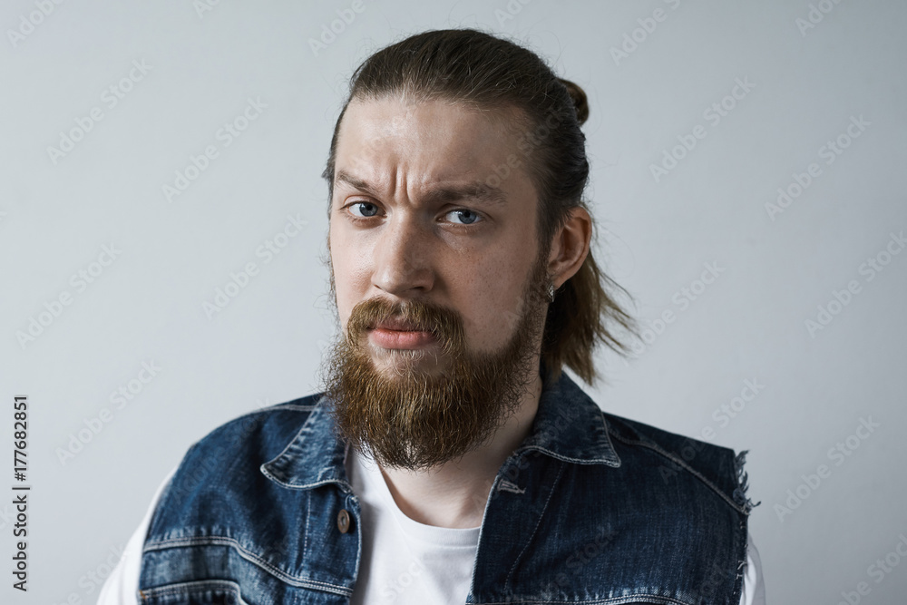 Displeased angry stylish young man with fuzzy beard, hair knot and earring staring at camera disapprovingly. Bearded guy in jeans vest frowning, expressing his dislike, disrespect or disapproval