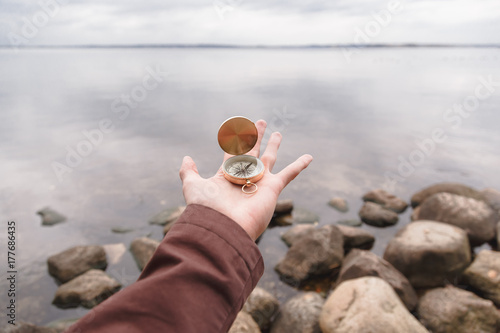 A traveler man holds a compass in his hand, standing on a stone beach. First person look