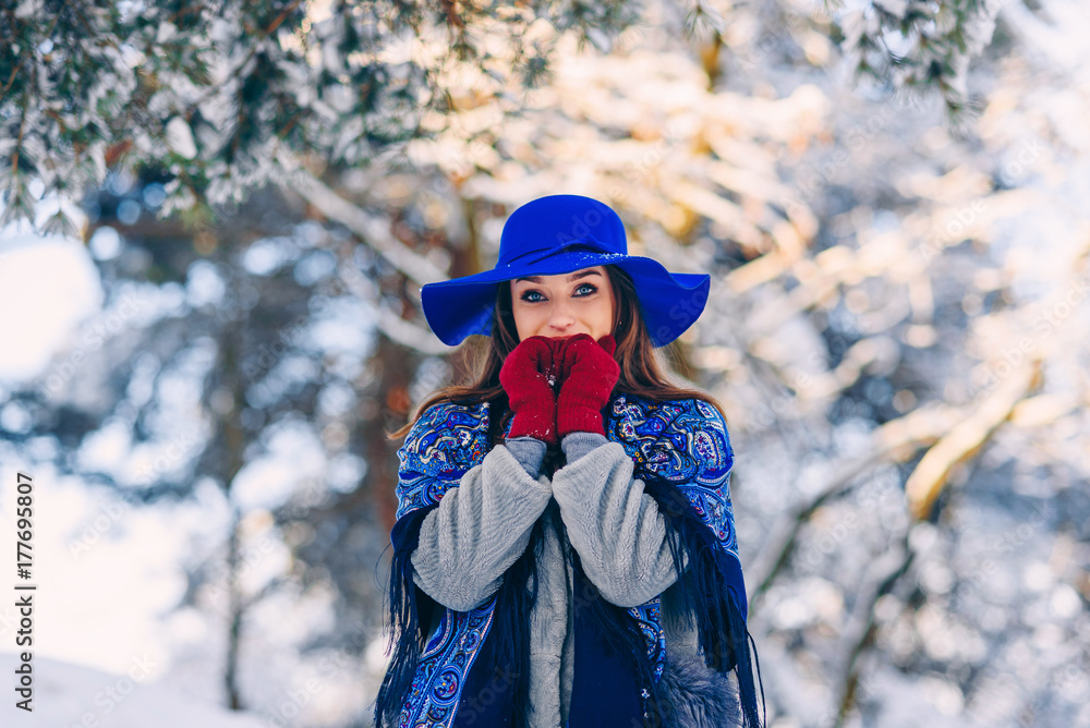 Young beautiful stylish girl in blue hat and scarf walking in the park on the background of snowy trees on sunset.