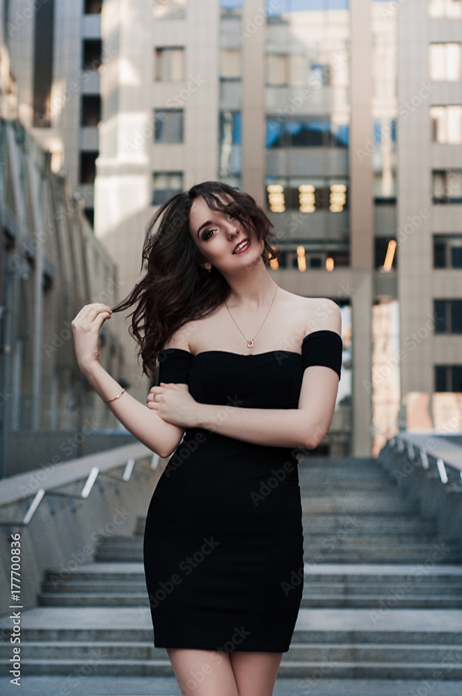 Outdoor lifestyle portrait of pretty young girl, wearing in black cocktail dress on urban background.Fashion street photo of stylish young lady/Picture of a young fashionable girl in the city
