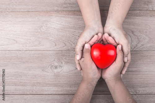 Love Mom Concept   Woman holds her young kids hands supporting red heart on brown wooden table. Free space for text of Mother s Day celebration.