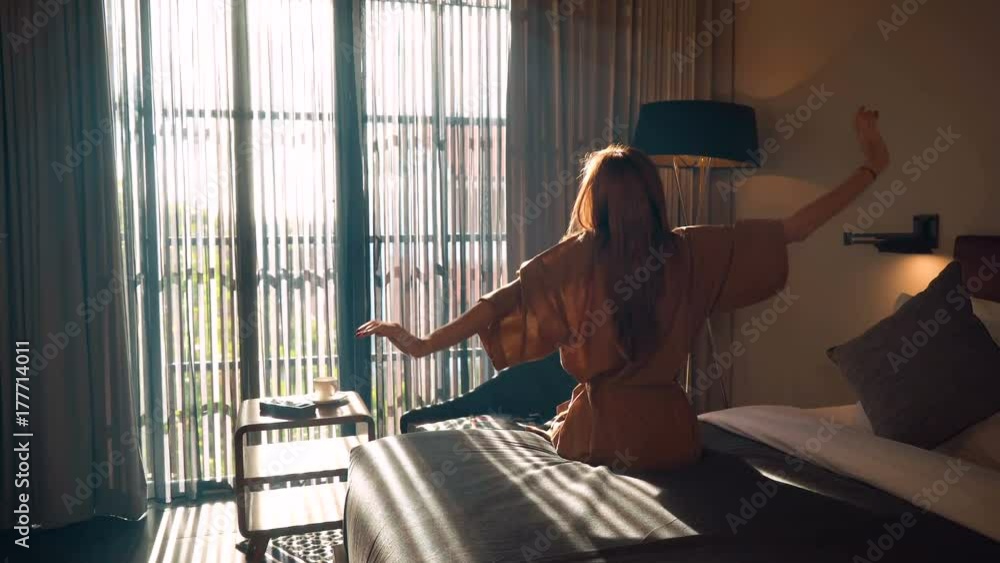 Young caucasian woman in brown beige robe stretches out hands sitting on neat bed in the morning in front of big window. Shot with Sony a7s and Atomos Ninja Flame.