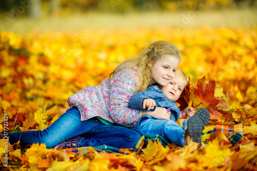 happy sister hugging her younger brother sitting in the park on fallen autumn foliage.