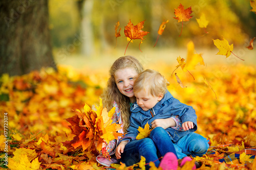 happy sister hugging her younger brother sitting in the park on fallen autumn foliage.