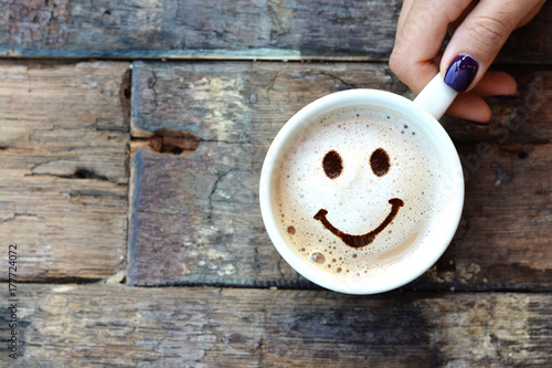 Murais de parede Happy face on cappuccino foam, woman hands holding one cappuccino cup on wooden