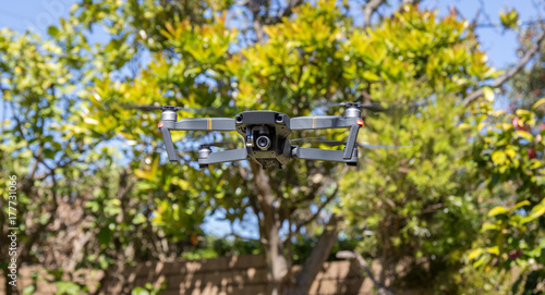 Small foldable drone flying viewed from below with green trees in the background