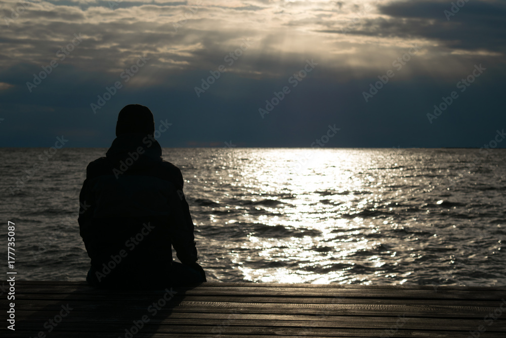 Silhouette of lonely man in jacket and cap who seating on wooden dock and looking on sunset over sea. Cold autumn.