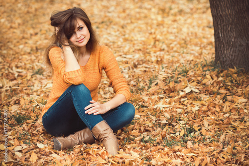 fashion portrait of a girl in jeans and a sweater in the nature sitting on yellow autumn leaves