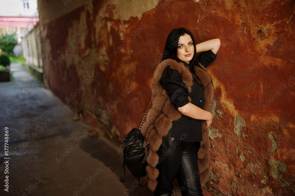 Fashion outdoor photo of gorgeous sensual woman with dark hair in elegant clothes and luxurious fur coat at old street with grunge walls.