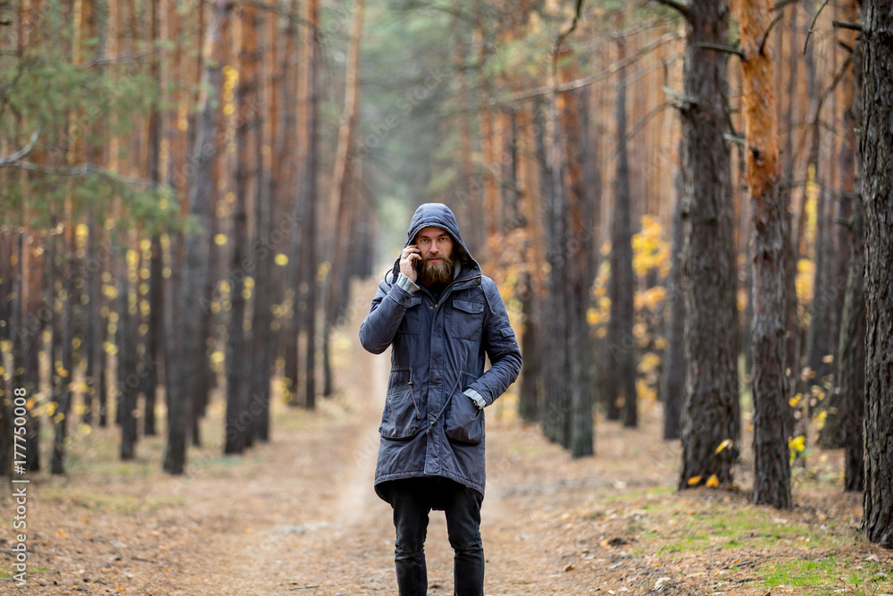 A man in a gray jacket stands on the road in the middle of a pine forest and talks on the phone