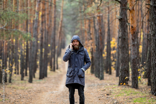 A man in a gray jacket stands on the road in the middle of a pine forest and talks on the phone © drotik