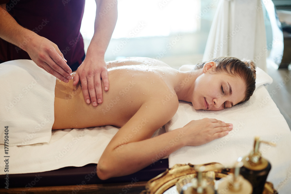 Fototapeta premium Portrait of blissful young woman enjoying massage in SPA center, lying on massage table with eyes closed and smiling with male masseur rubbing her back