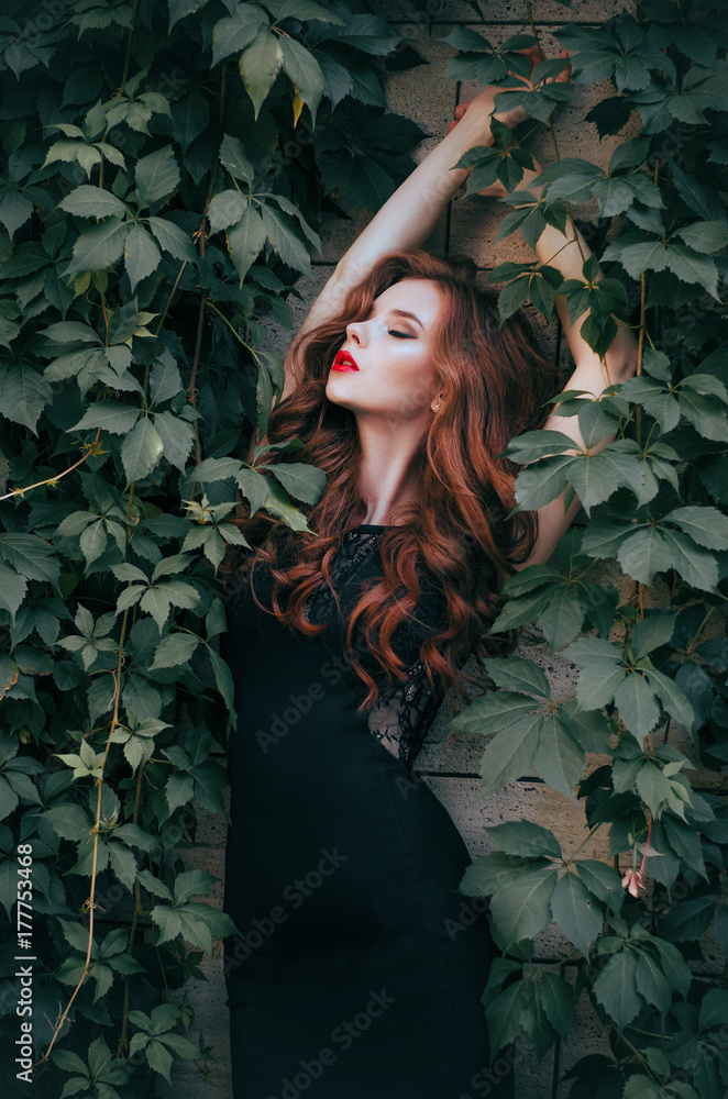 fashion model with red hair in a long black dress posing against a wall  with green leaves. A white-skinned redhead with red lips. Fashion photo.  Stock Photo | Adobe Stock