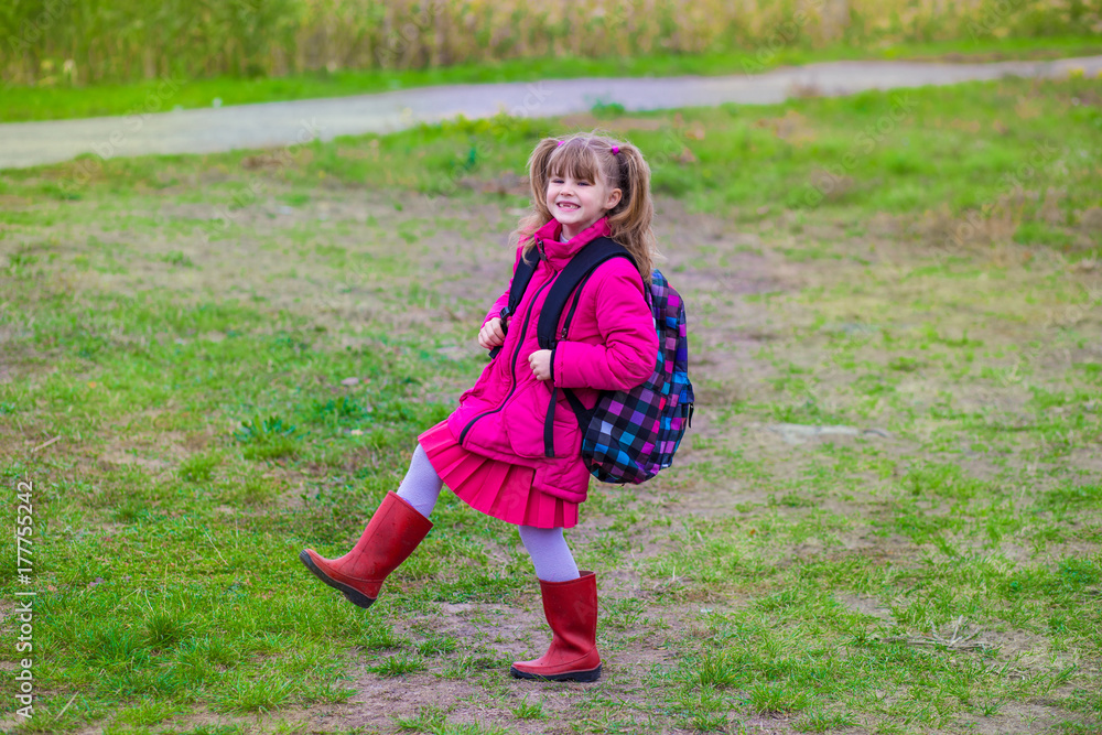 Fototapeta premium Little cute schoolgirl with a big backpack walking on the grass in the autumn day