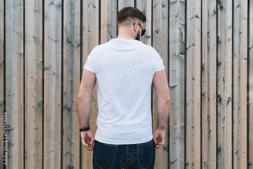 Summer day. Back view. Young bearded hipster man dressed in white t-shirt and sunglasses is stands outdoor against wood wall. Mock up. Space for logo, text, image.