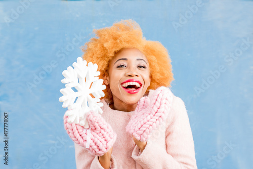 Portrait of a cute african woman in the pink coat holding an artificial snowflake on the blue wall background. Winter season concept